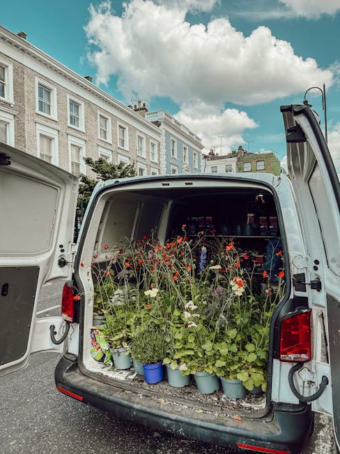 The rear of a white delivery van parked on a street with its doors open, revealing an interior filled with potted plants and fresh flowers, including bright red poppies, white daisies, and green folia